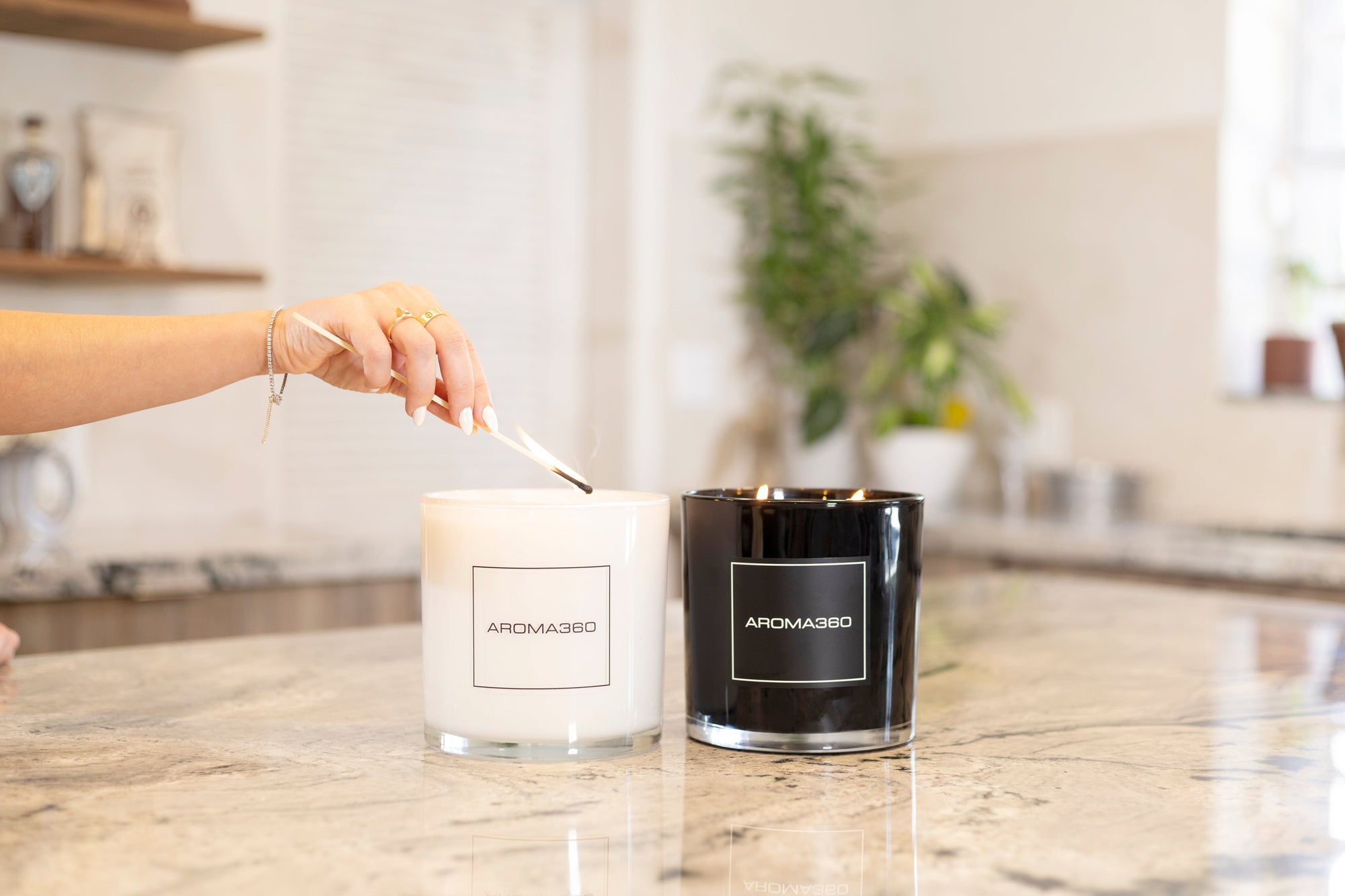 A hand is lighting a white Aroma360 4-wick deluxe candle using a matchstick, with a black version of the same candle already lit next to it on a marble countertop in a modern kitchen setting.